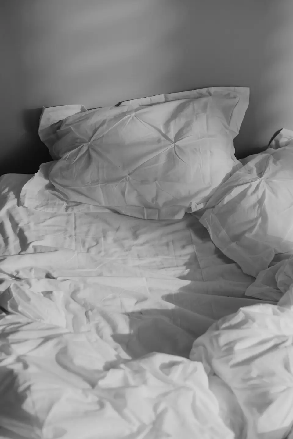 Crumpled white pillows and bedsheets lie across an unmade bed in soft natural light.