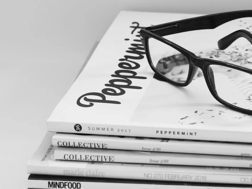 A pair of black-framed glasses rests on top of a stack of glossy magazines on a white surface.