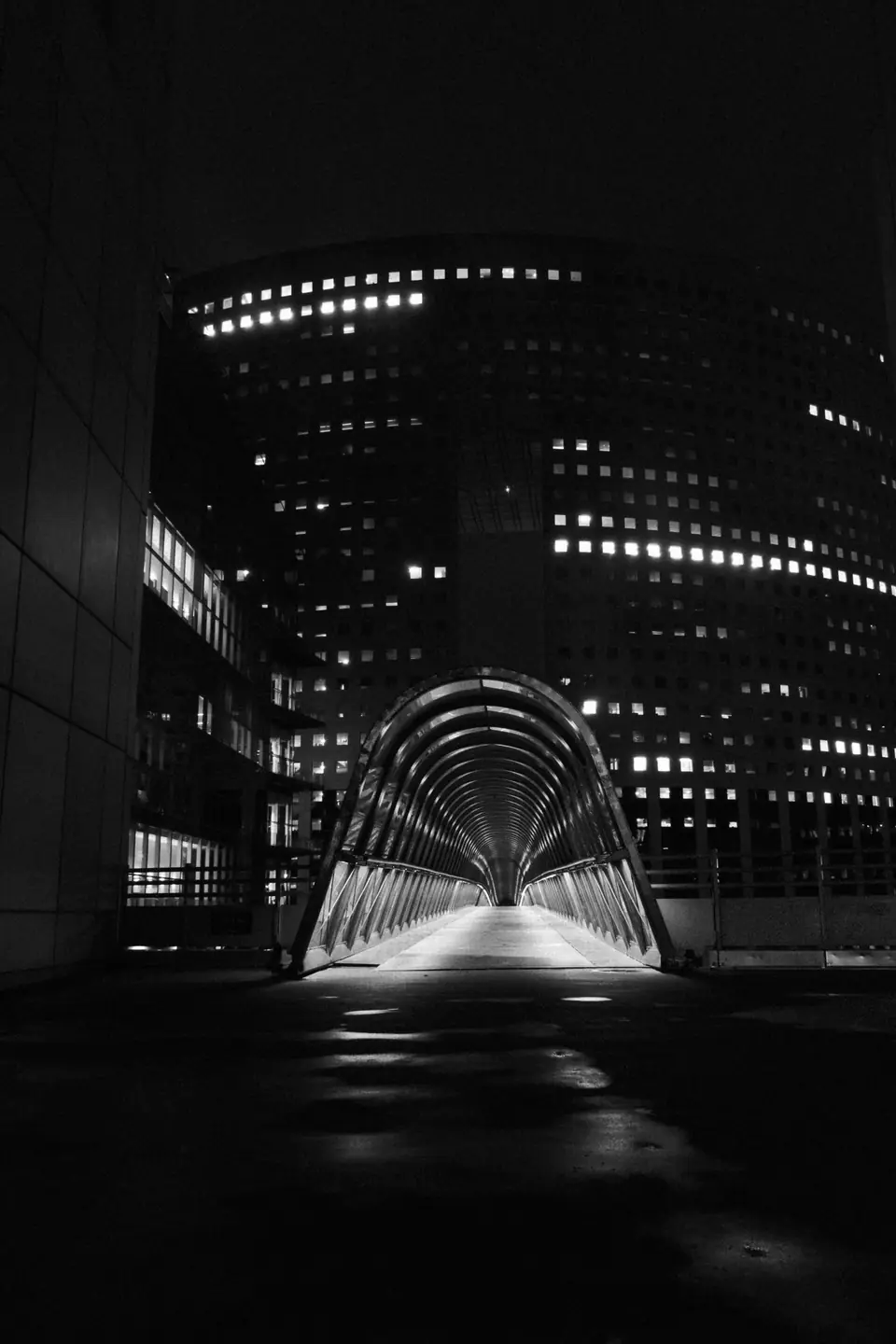 A lighted, tunnel-like pedestrian bridge leads toward a tall office building with lit windows at night.