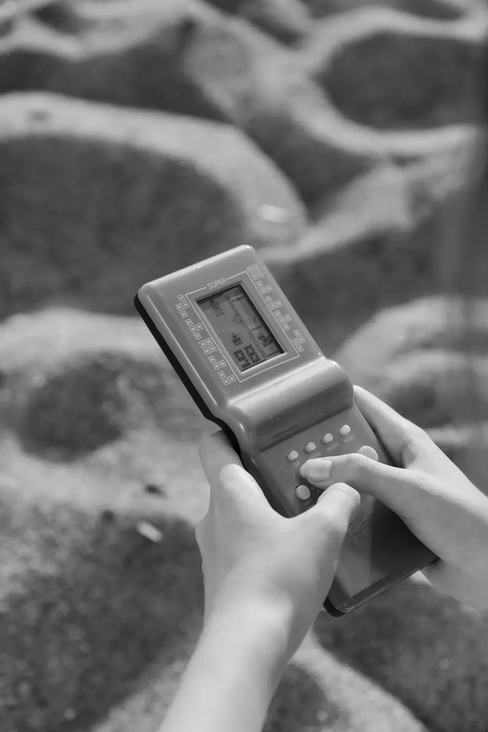 Hands hold a small handheld brick game with a monochrome screen above a sandy surface.