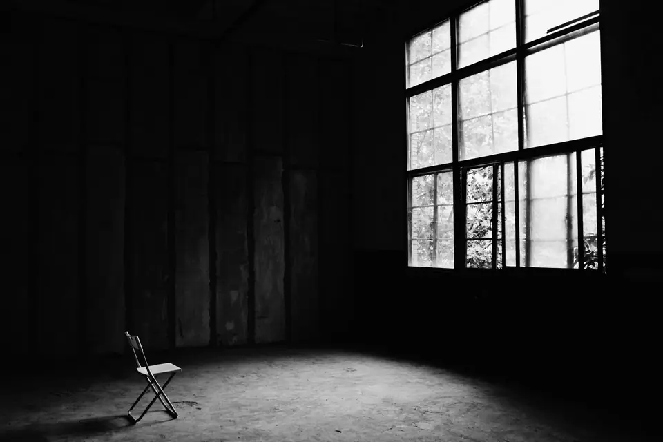 A lone folding chair stands on the floor of a large, dark, empty room beside a bright window.