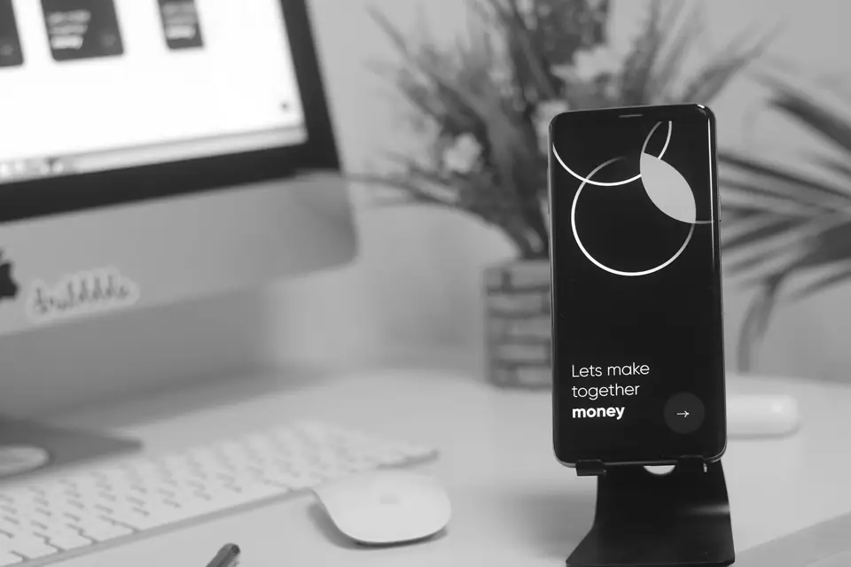 A smartphone on a black stand displays a promotional screen in front of a monitor, keyboard and potted plant.