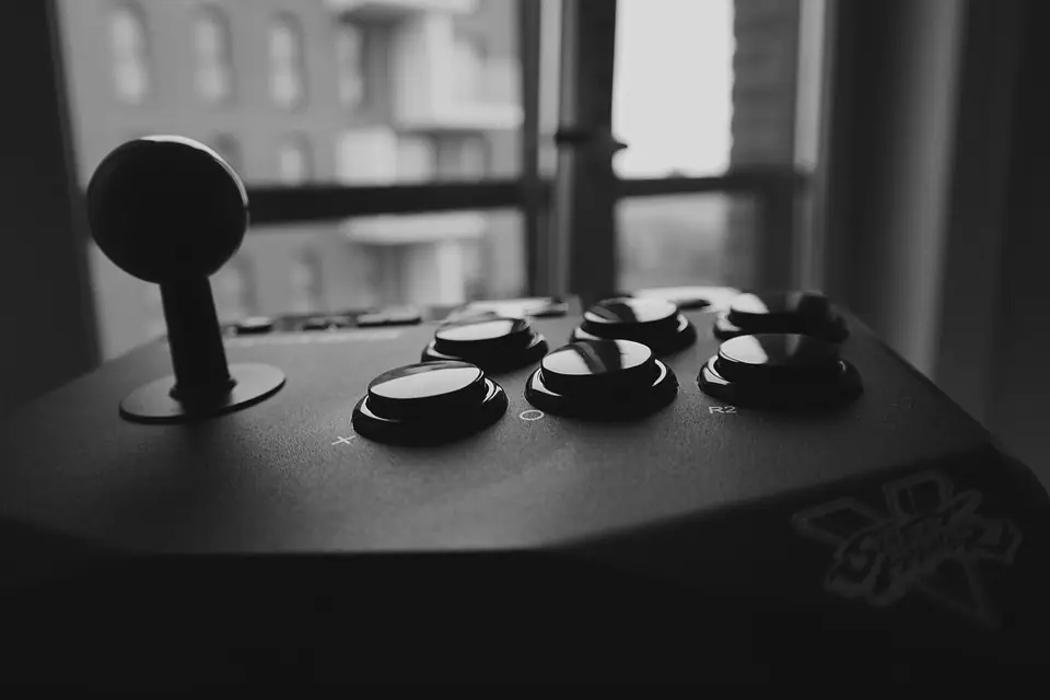 A black arcade joystick and round buttons on a controller sit near a bright window.
