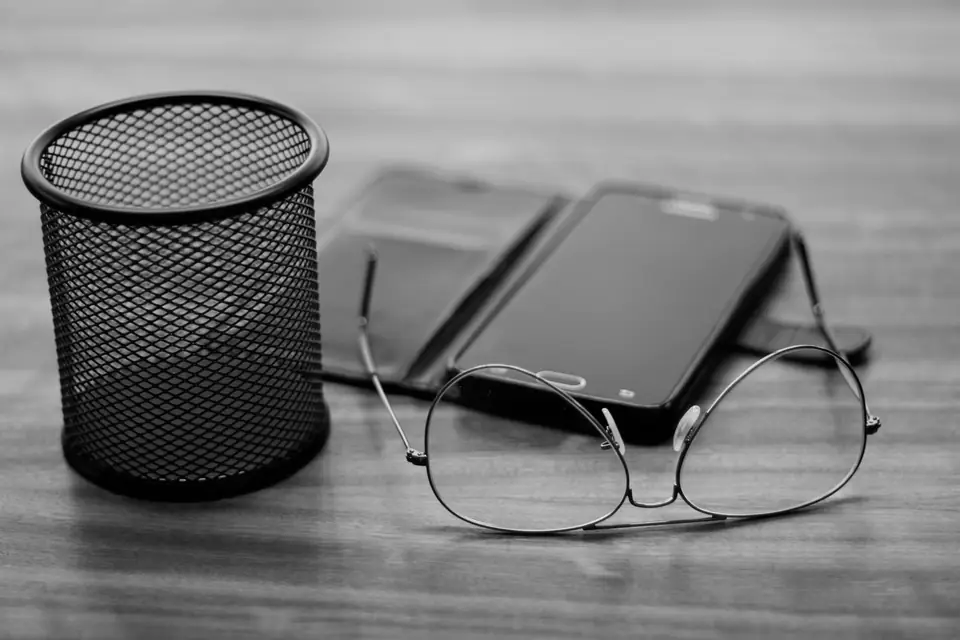 Two smartphones lie on a wooden desk beside a pair of glasses and a mesh pencil cup.