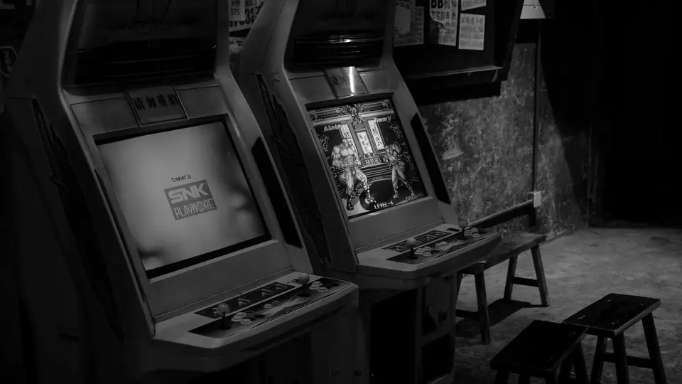 Two arcade cabinets with lit screens stand side by side in a dim room with stools.