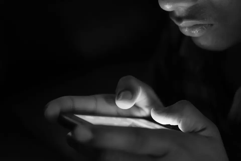 A person’s hands hold a glowing smartphone in the dark, the lower half of their face barely visible.