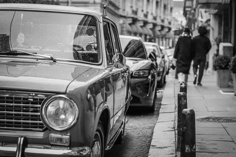 A small vintage sedan is parked along a city street as two people walk down the sidewalk.