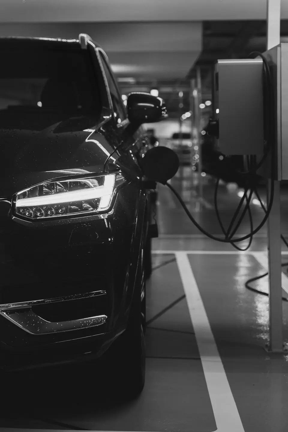 A dark SUV is connected by a charging cable to a wall-mounted charger in an indoor parking garage.