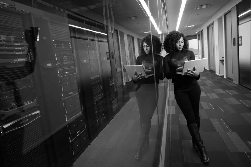 A woman holding a tablet leans against a glass wall beside rows of server racks in a corridor.