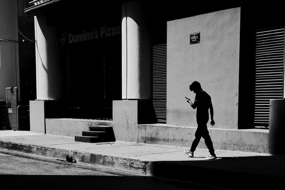 A man in silhouette walks past a storefront while looking at his phone, under a legible “Domino’s Pizza” sign.