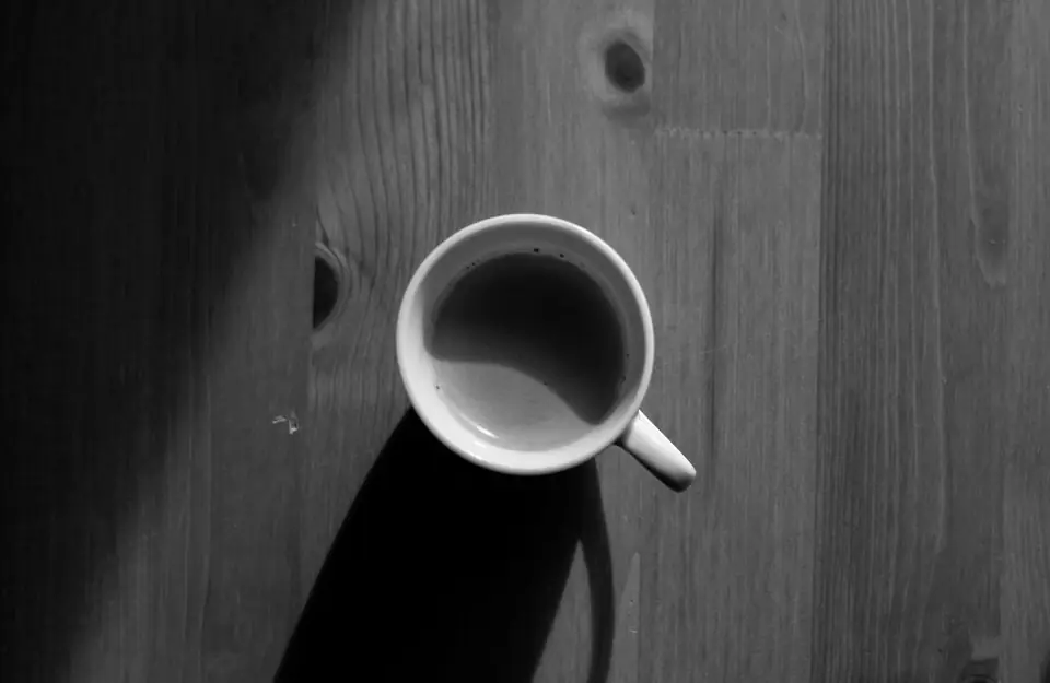 A small white cup of coffee sits on a wooden table, seen from directly above.