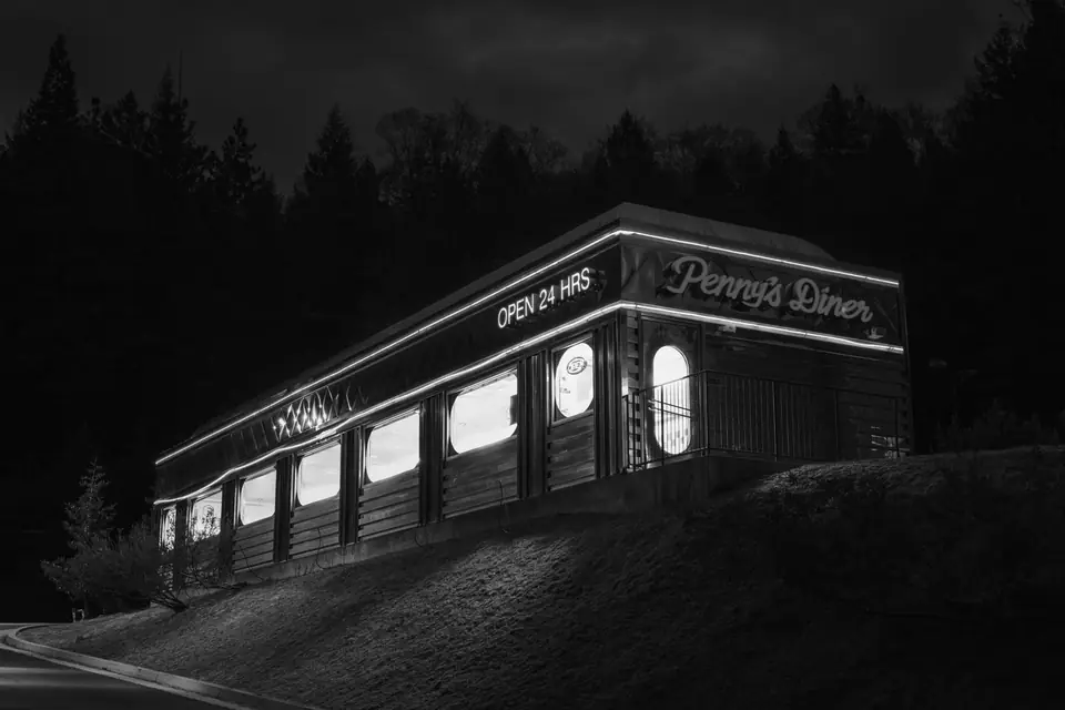 A diner with a sign reading “Penny’s Diner” and “Open 24 Hrs” glows at night below dark trees.