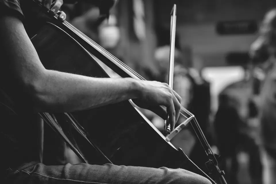 A seated musician draws a bow across a cello, with other people blurred in the background.
