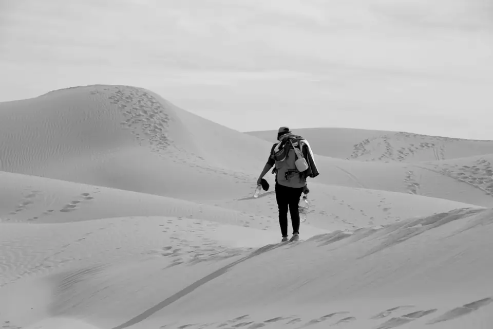 A person carrying a backpack walks up a sand dune marked with footprints.