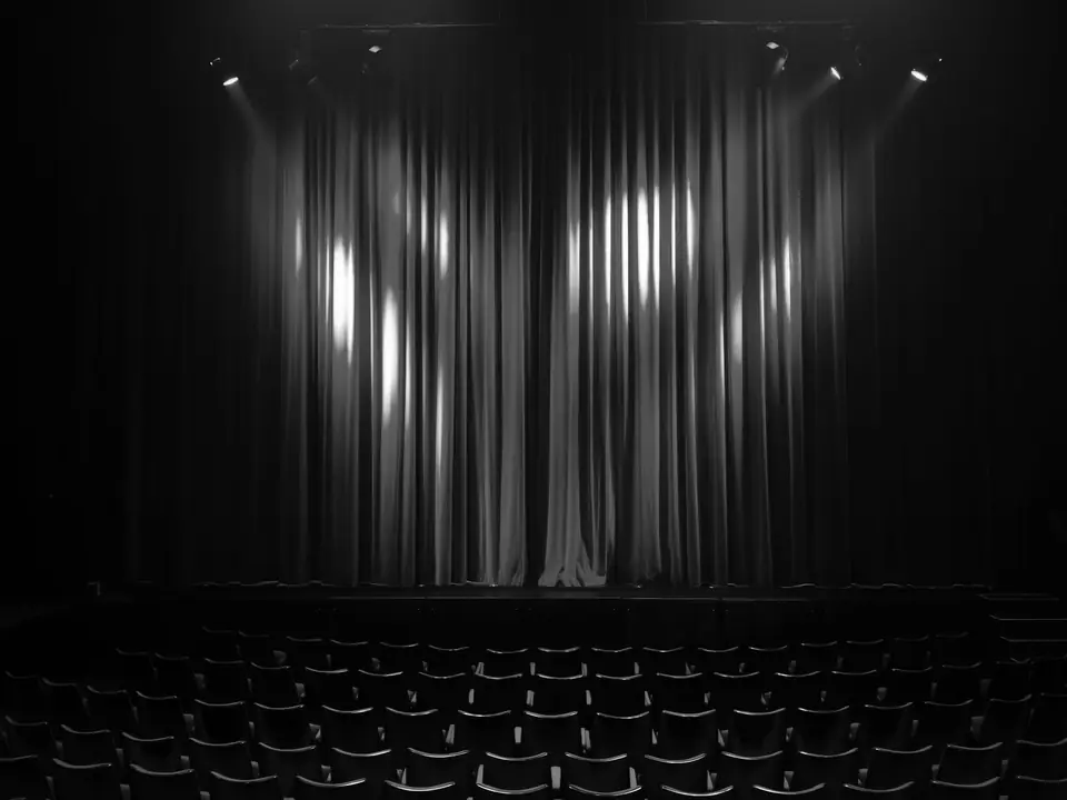A dark theater with rows of empty seats faces a closed stage curtain lit by spotlights.