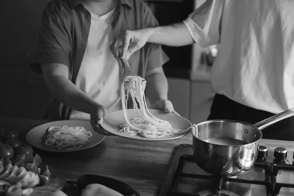 One person twirls spaghetti onto a plate held by another person beside a saucepan on a stove.