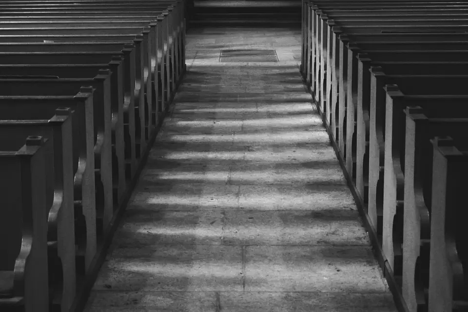 A stone aisle runs between two long rows of wooden church pews in soft light.