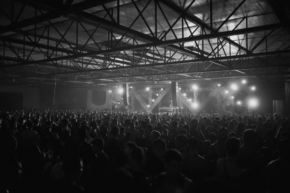A large crowd fills a warehouse venue facing a brightly lit stage under steel roof trusses.