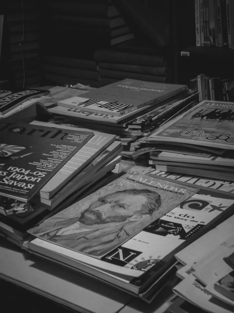 Stacks of old magazines with worn covers lie piled across a table in a dimly lit room.