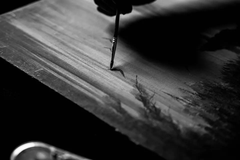 A thin paintbrush adds fine detail to a painted landscape on a wooden panel.