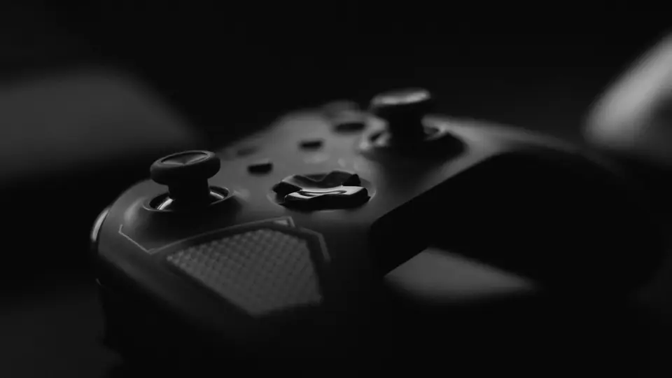 A dark game controller with thumbsticks and a textured grip sits mostly in shadow.