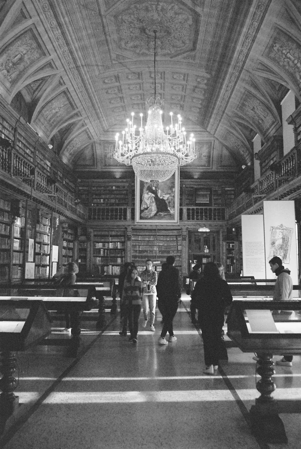 People walk through a long, ornate library hall with reading tables, tall shelves and a hanging chandelier.