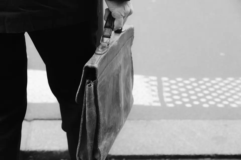 A person in dark trousers carries a scuffed leather briefcase beside a street curb with tactile paving.