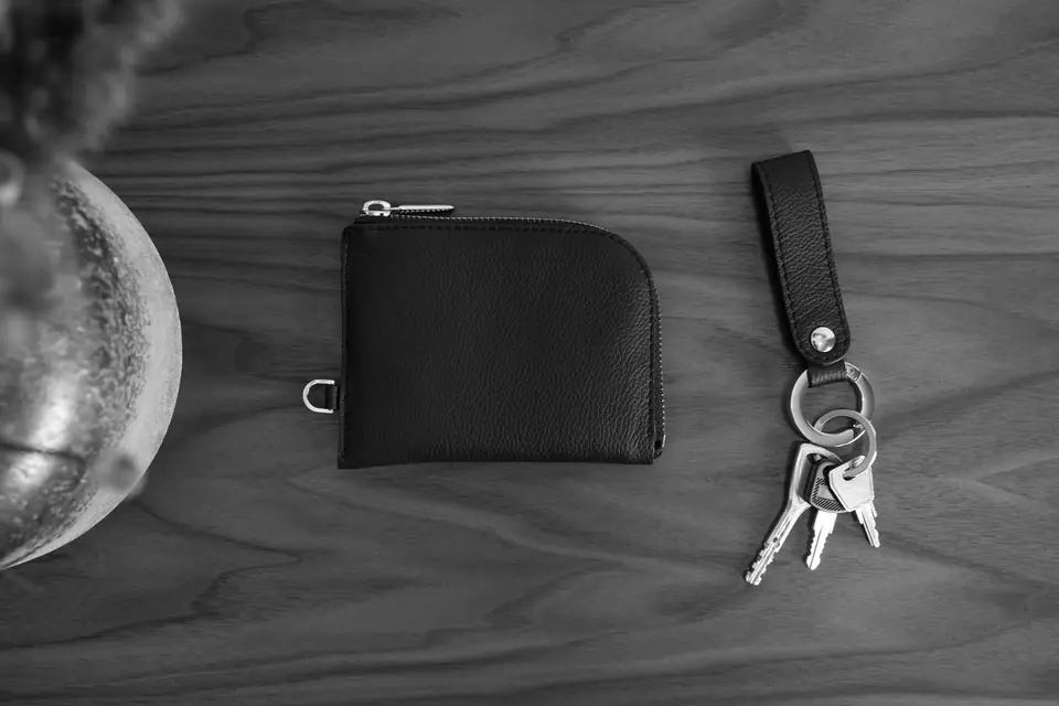 A small black zip wallet and a set of keys on a leather fob lie on a wooden tabletop.