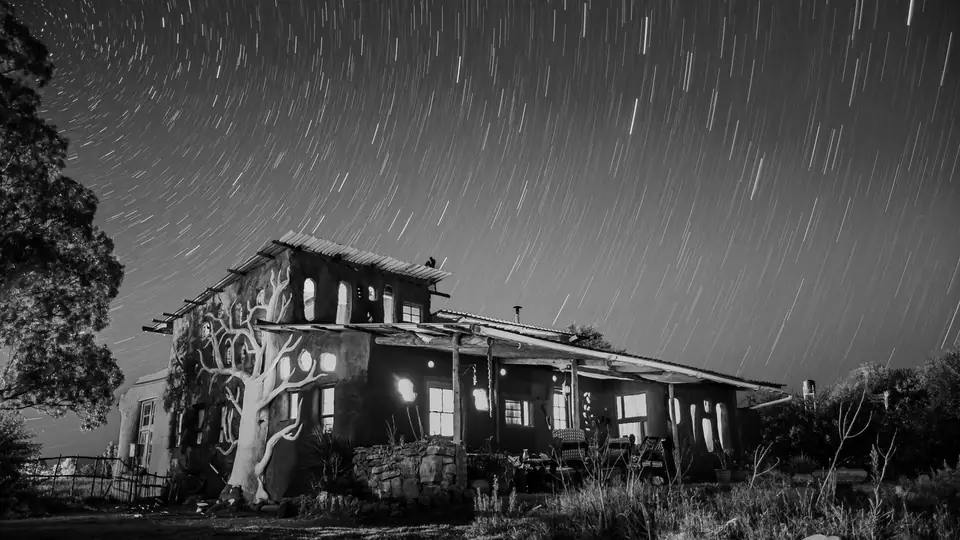 A two-story house with lit windows stands under circular star trails in a night sky.