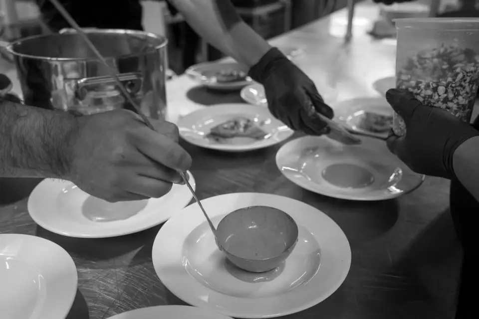 Several hands ladle soup from a large pot into white bowls and plates set out on a table.