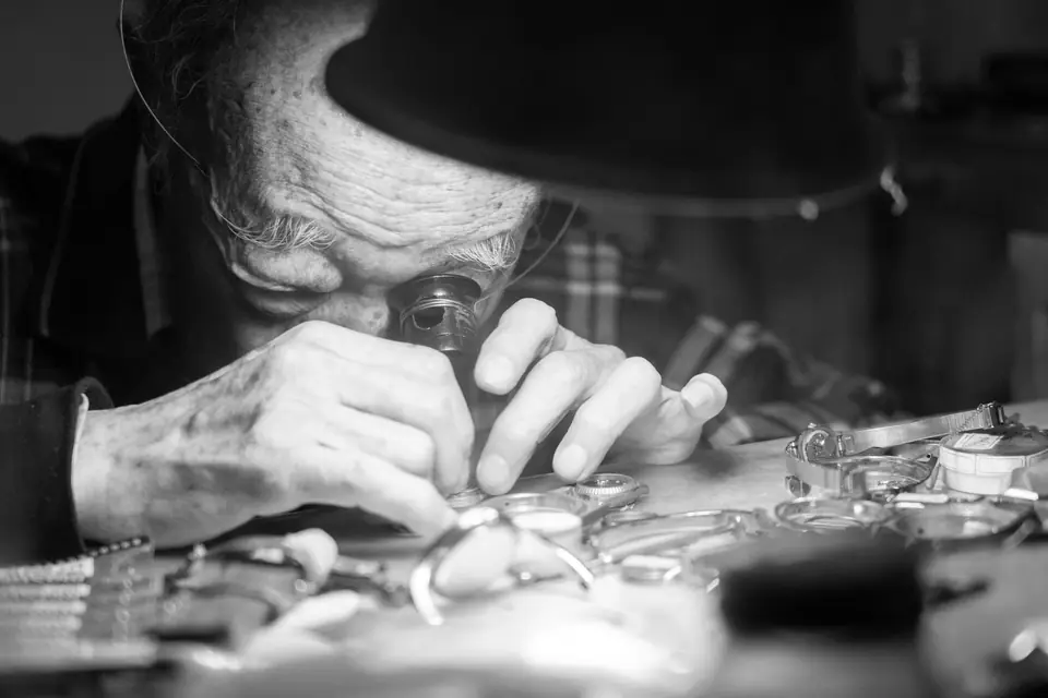 An elderly watchmaker wearing a loupe and a hat works with tweezers over a bench crowded with watch parts.