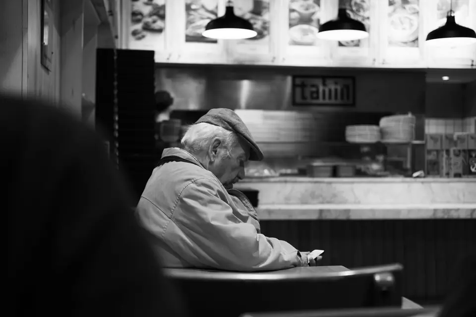 An older man in a cap and jacket sits alone in a restaurant booth, looking down.