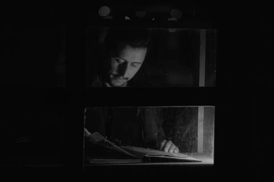 A man’s face and a book on a table are dimly lit through window openings in a dark room.