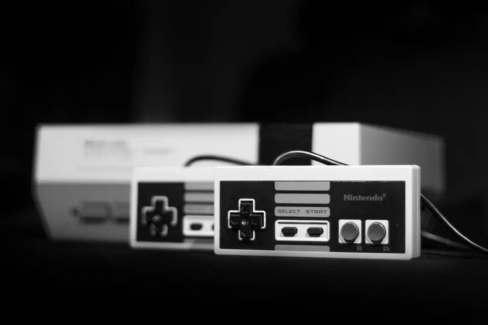 A gray retro game console sits behind two rectangular controllers against a dark background.