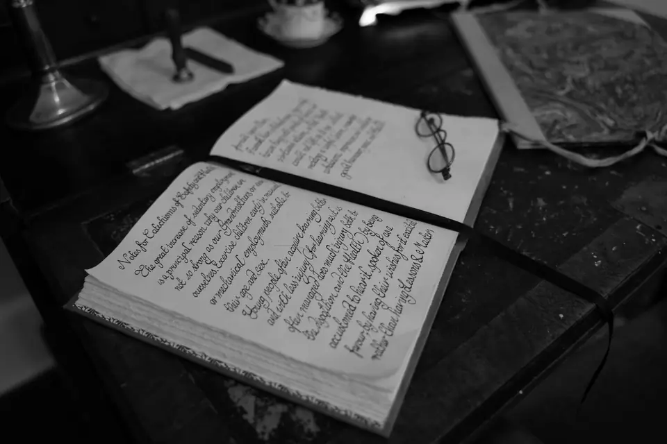 An open handwritten book and a pair of eyeglasses lie on a dark, cluttered desk.
