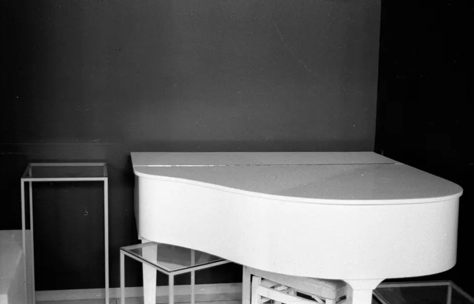 A white grand piano stands in a dark, sparsely furnished room beside glass tables.