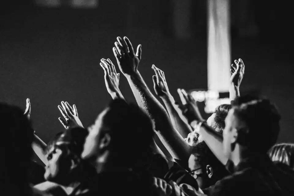 A dense crowd of young people raise their hands toward a dark stage at a concert.