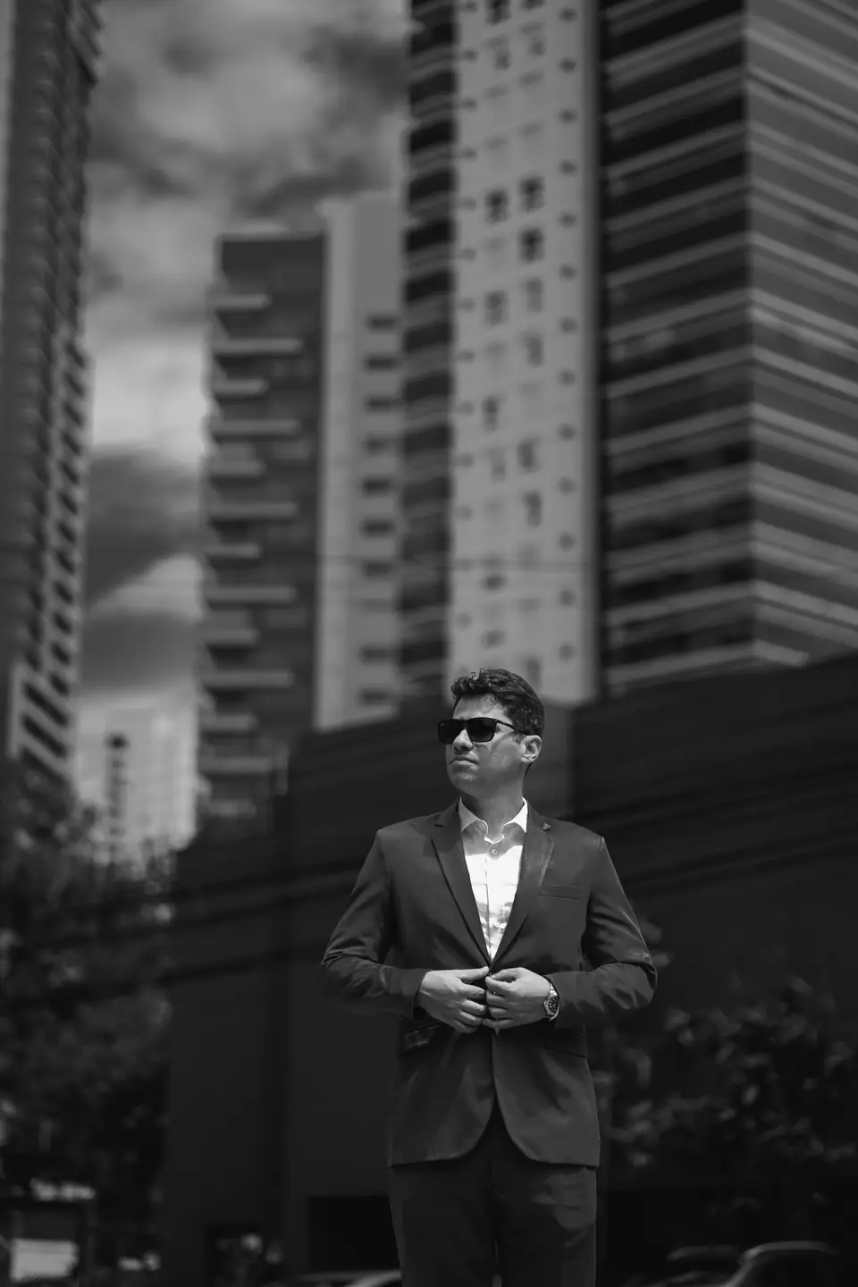 A man in sunglasses and a dark suit buttons his jacket in front of tall apartment towers.