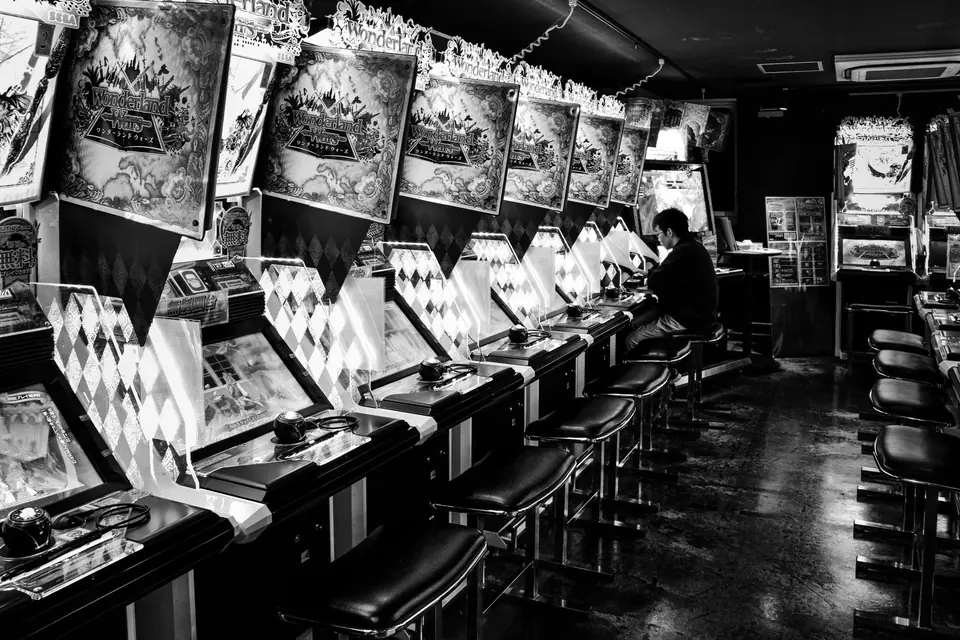 A long row of arcade machines with stools lines a dark room where one person sits playing.