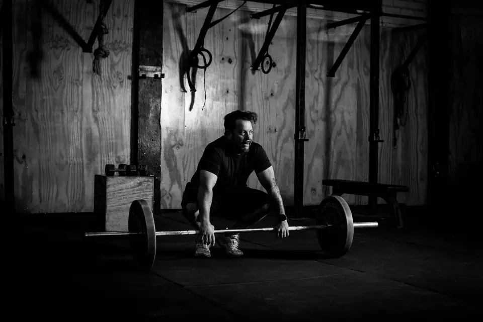 A man crouches beside a loaded barbell on the floor of a dim gym with wooden walls.