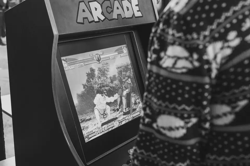A person in a patterned sweater stands beside an arcade cabinet labeled “Arcade” showing a fighting game.