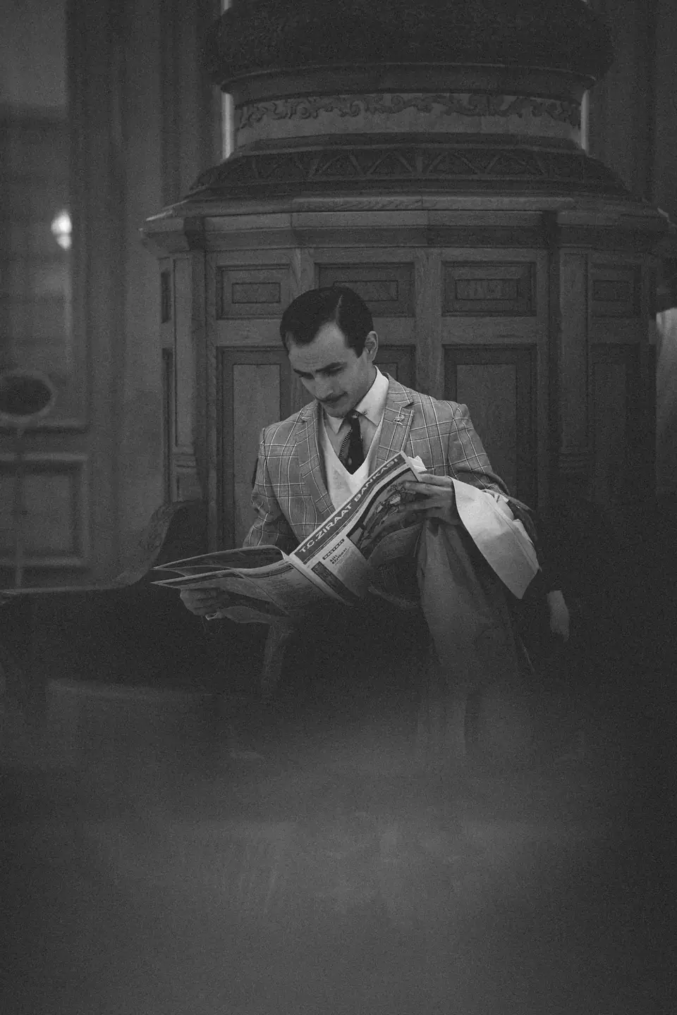 A man in a checked suit and tie reads a newspaper in a dark, wood-panelled interior.