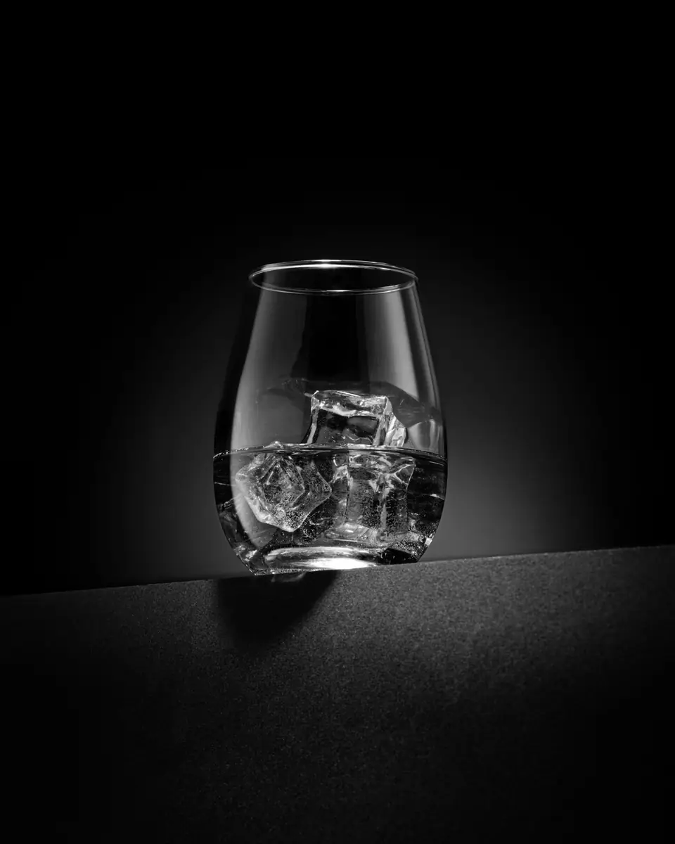 A stemless glass holding ice cubes and a small amount of clear liquid sits in a spotlight on black.