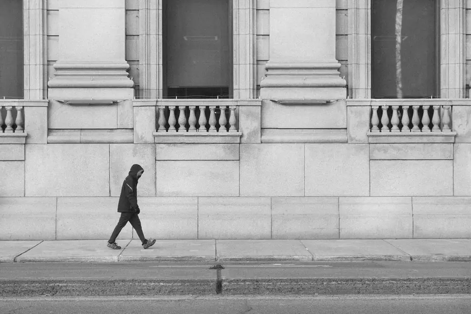 A person in a dark hooded jacket walks along a sidewalk in front of a long stone building.