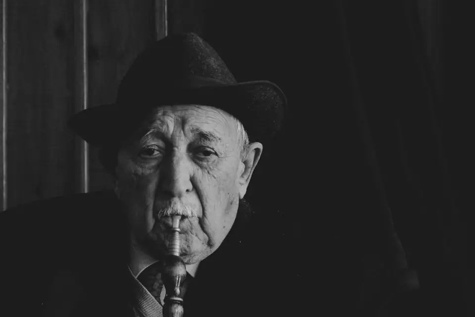 An elderly man in a dark hat and jacket, holding a pipe in his mouth, looks at the camera.