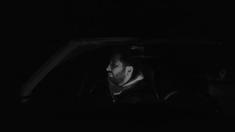 A bearded man sits in the driver’s seat of a car at night, dimly lit.