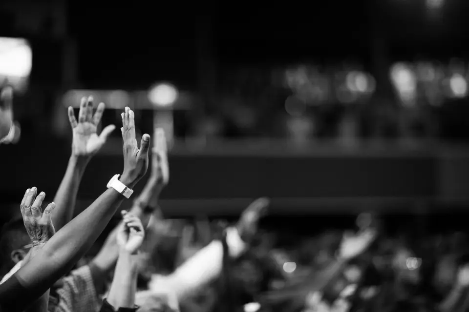 Several raised arms, one with a wristwatch, reach up above a blurred concert crowd.