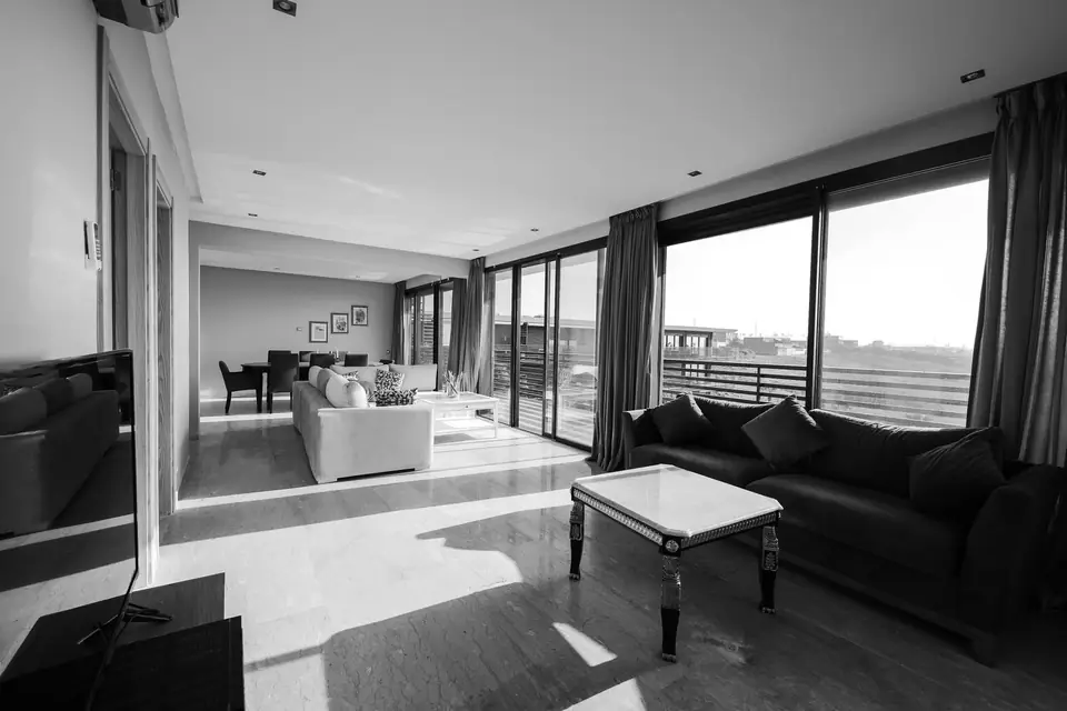 A spacious living room with dark sofas, a white coffee table and floor-to-ceiling windows overlooks a balcony.