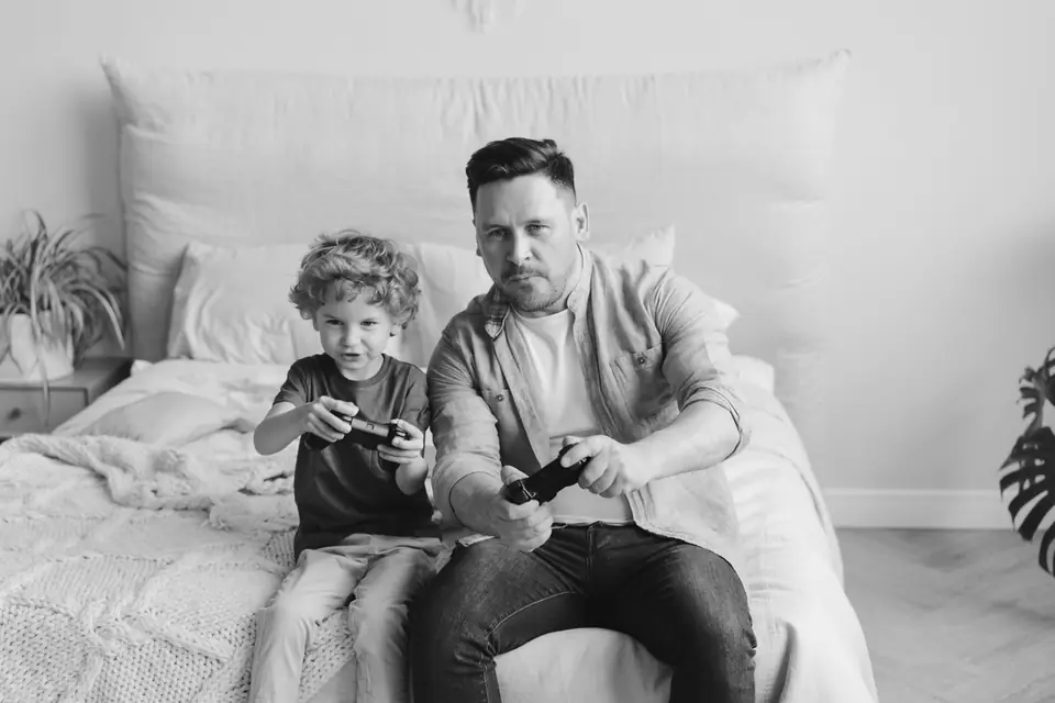 A man and a small curly-haired boy sit on a bed holding game controllers.