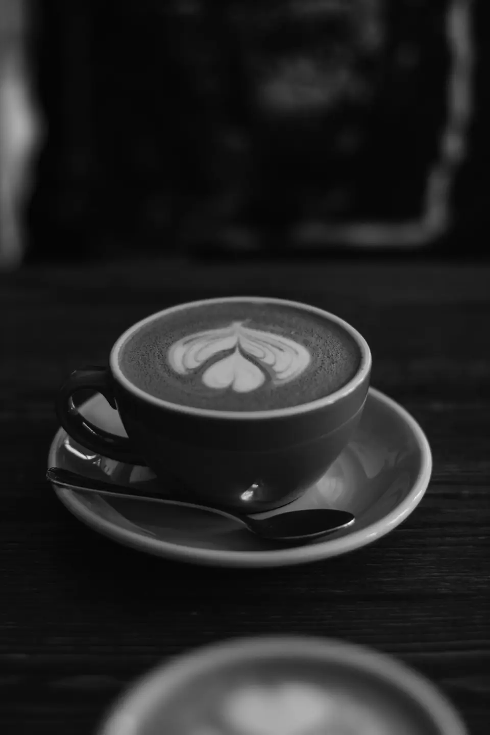 A cup of coffee with latte art in its foam rests on a saucer with a spoon on a dark table.